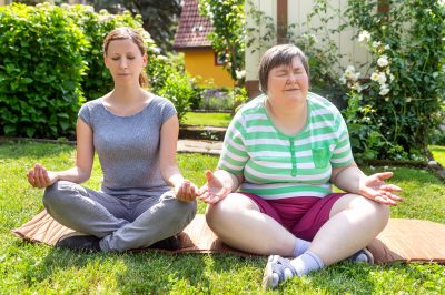 two women, one of them a fitness or yoga coach, are doing some yoga or relaxation exercises, on woman is mentally disabled